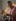 Ben W in a pink short‑sleeve button‑up shirt preparing food in a kitchen, placing fresh herbs into a glass jar near a stovetop with light-colored cabinets and a tiled backsplash.