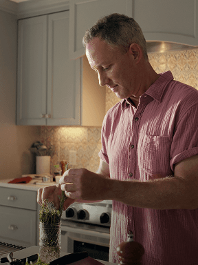 Ben W in a pink short‑sleeve button‑up shirt preparing food in a kitchen, placing fresh herbs into a glass jar near a stovetop with light-colored cabinets and a tiled backsplash.