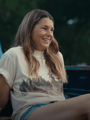 Jessica S sits outdoors at the back of a vehicle, wearing a light-colored T‑shirt with an illustration of horses and denim shorts, with trees visible in the background.