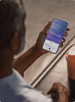 A person holding a smartphone displaying the Lingo app with a glucose reading, seated at a table next to a shaker bottle.