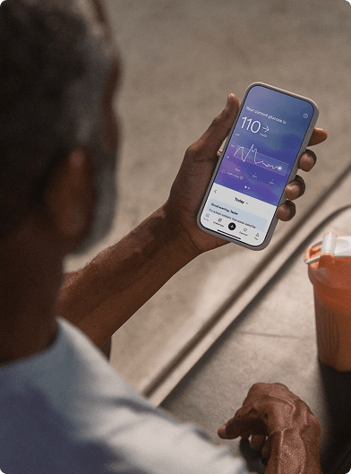 A person holding a smartphone displaying the Lingo app with a glucose reading, seated at a table next to a shaker bottle.