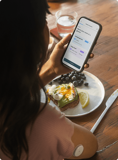 A person seated at a wooden table holding a smartphone displaying the Lingo app, with a plate of food including toast, eggs, and fruit in front of them and a Lingo biosensor on the upper arm.