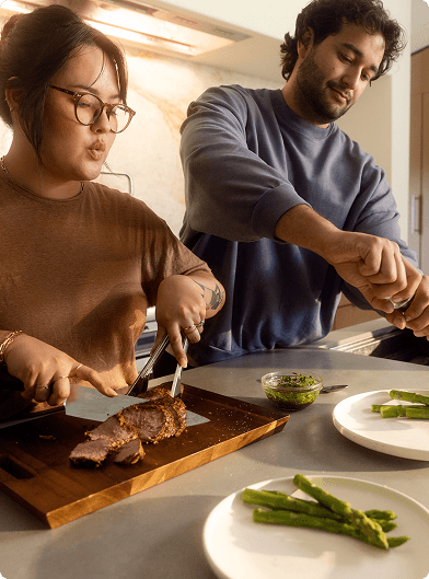 Two people preparing a meal in a kitchen; one is slicing cooked meat on a wooden cutting board while the other is seasoning food, with plates of asparagus and a small bowl of sauce on the counter.