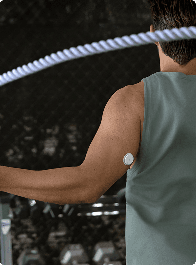 A person using a jump rope during a workout, viewed from behind, wearing a sleeveless top with a small round sensor attached to the upper arm. A row of weights is visible in the background.