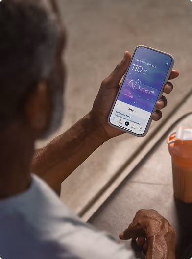 A person holding a smartphone displaying the Lingo app with a glucose reading, seated at a table next to a shaker bottle.