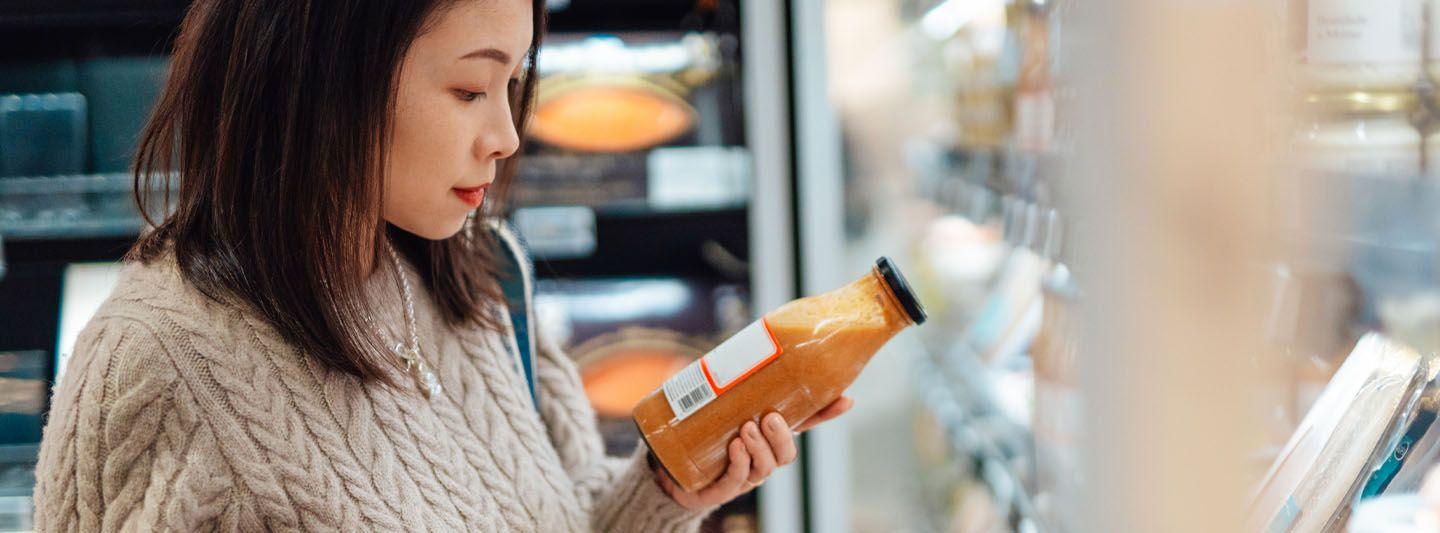 A person in a beige sweater holds a bottle of sauce while shopping in a grocery store aisle. Shelves with various items are visible in the background.