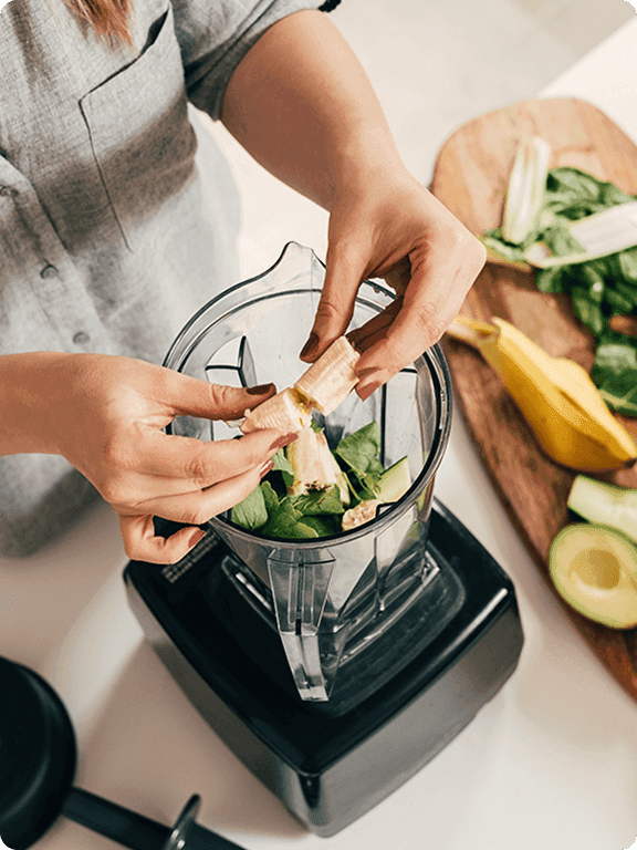 A photo of a person putting a banana into a blender that has other vegetables