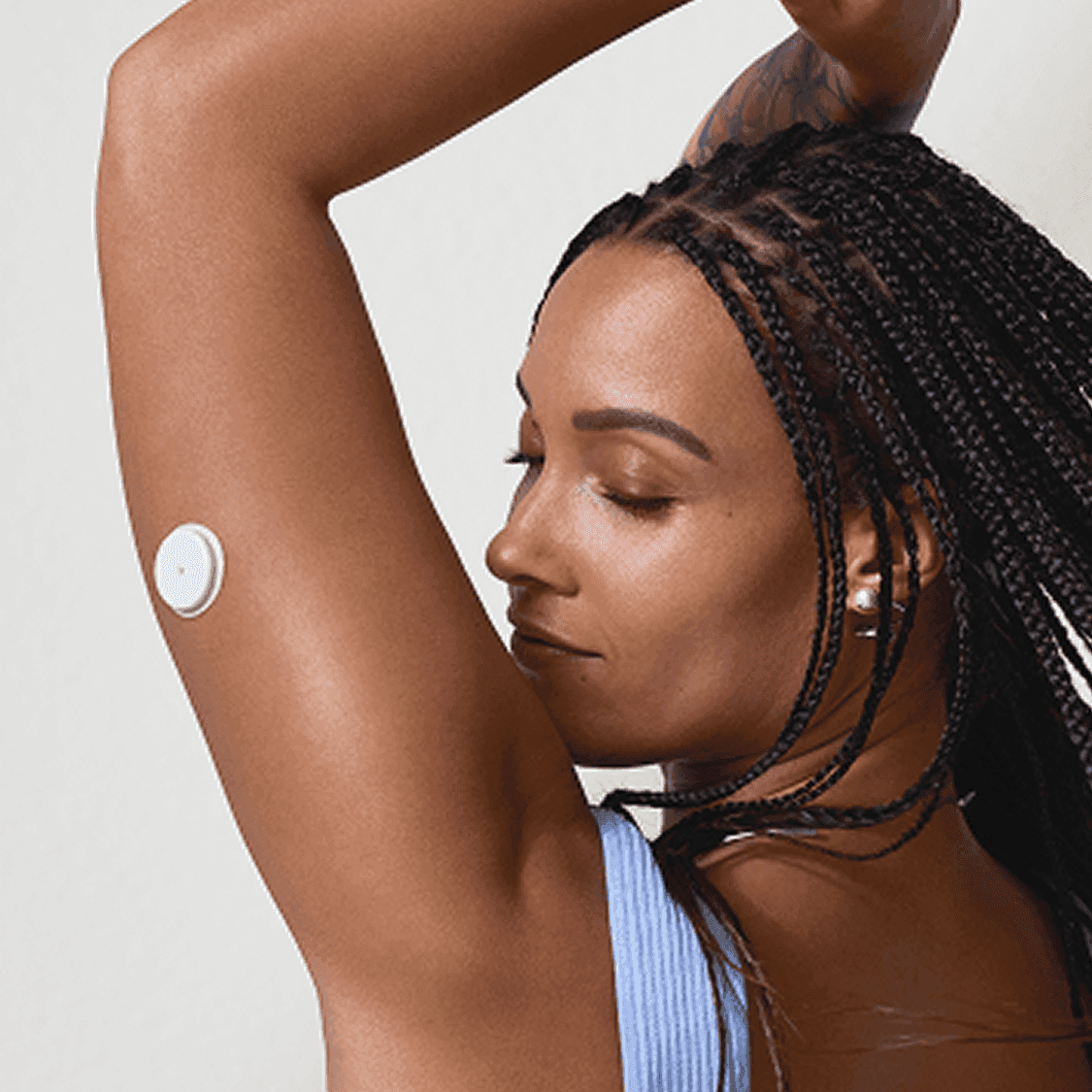 A person with braided hair, a tattoo on the right arm, and a circular medical device on the upper left arm, wearing a white sleeveless top. The background is a blue sky with clouds.