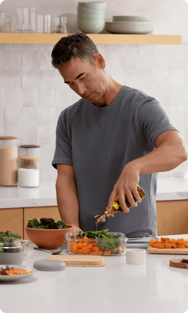 A person in a bright kitchen surrounded by prepped vegetables, pouring olive oil over a container of carrots and leafy greens.