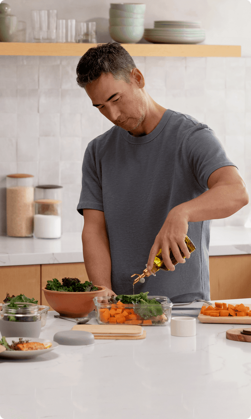 A person in a bright kitchen surrounded by prepped vegetables, pouring olive oil over a container of carrots and leafy greens.