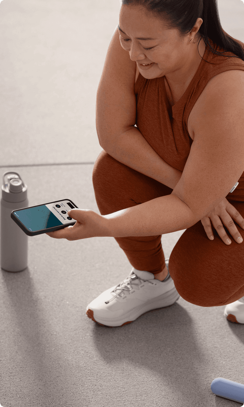 A person with a long ponytail wearing an earthy orange workout outfit, kneeling and smiling at a phone. They have a Lingo biosensor on their left arm.