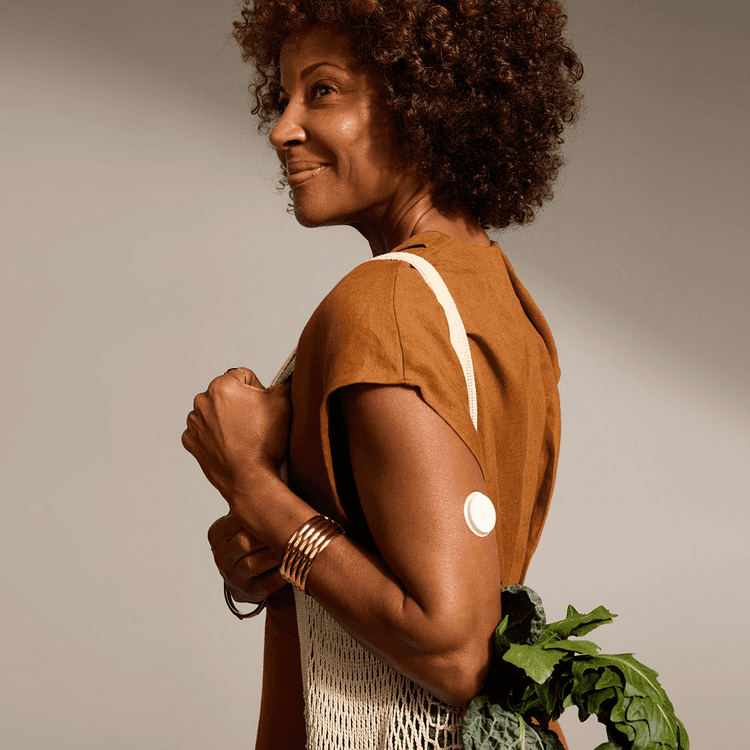 A smiling person with curly hair, wearing a Lingo biosensor, holding a macrame bag of leafy greens