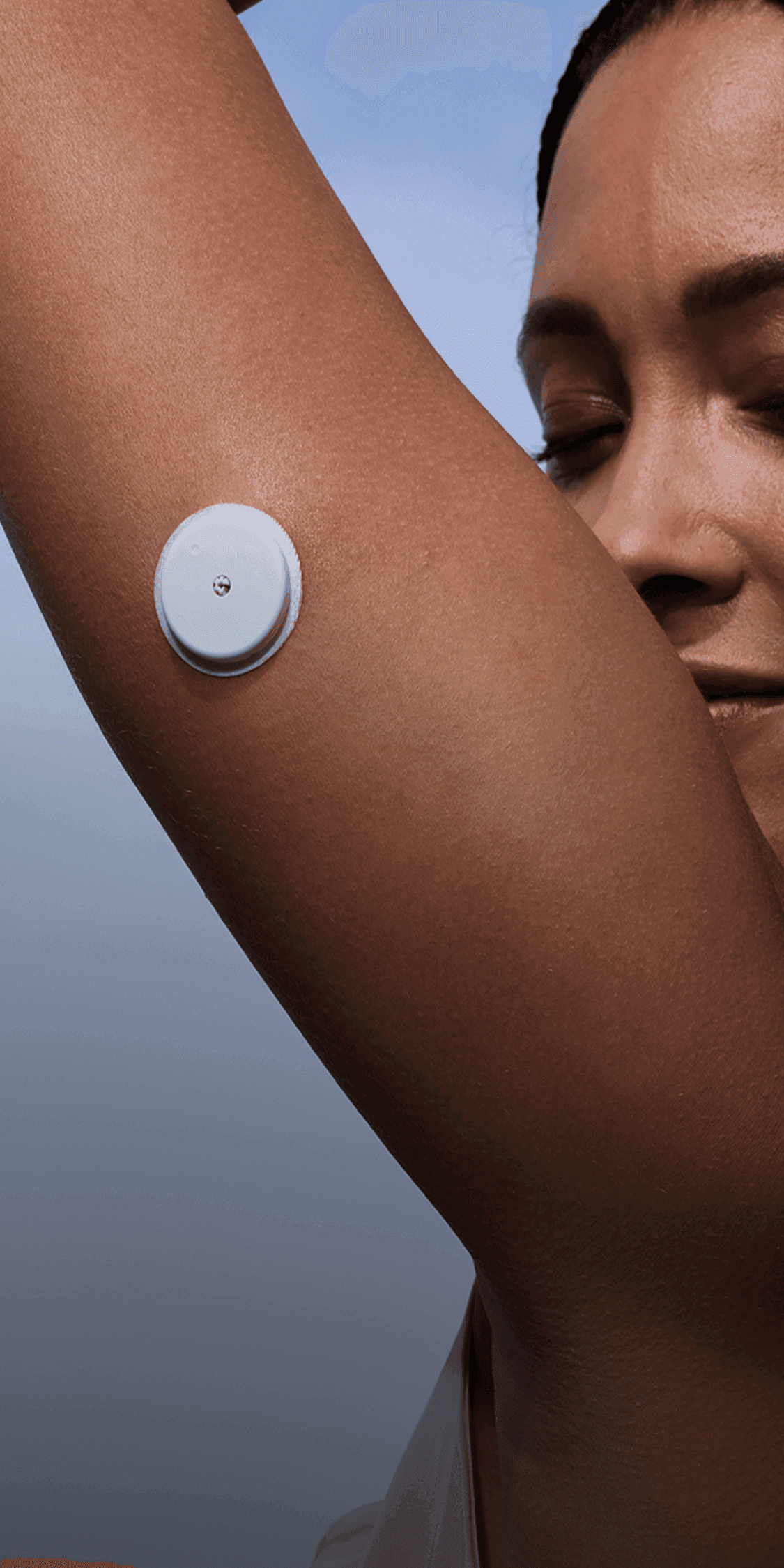 A person with braided hair, a tattoo on the right arm, and a circular medical device on the upper left arm, wearing a white sleeveless top. The background is a blue sky with clouds.